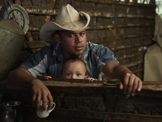 father with hat holding a cup of cofee and his son hiding behind 