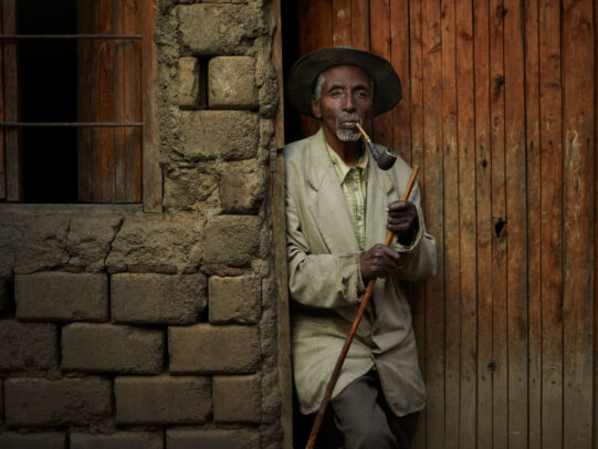 man with hat leaning on a door with a pipe and a stick