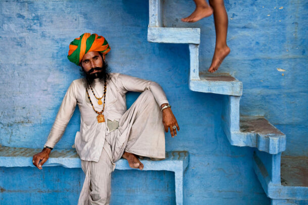 A man with orange and green turban sits beneath blue stairs