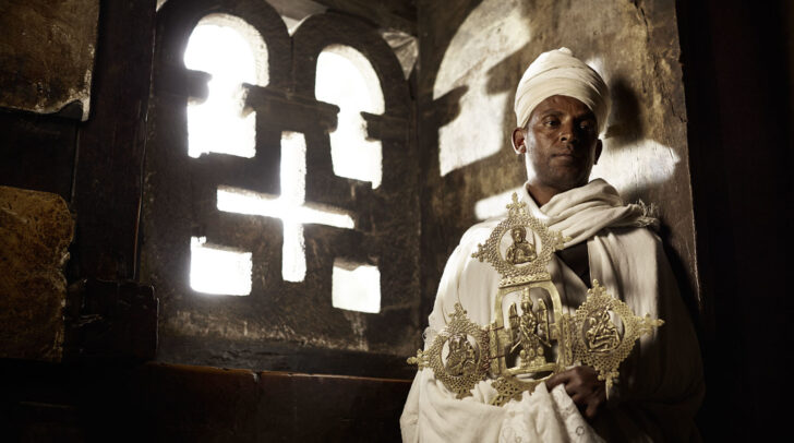 man wearing white robes and turban and holding a big golden cross near a crossed window