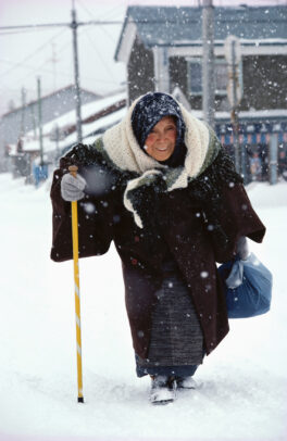 old woman walking on a snowy street with a stick